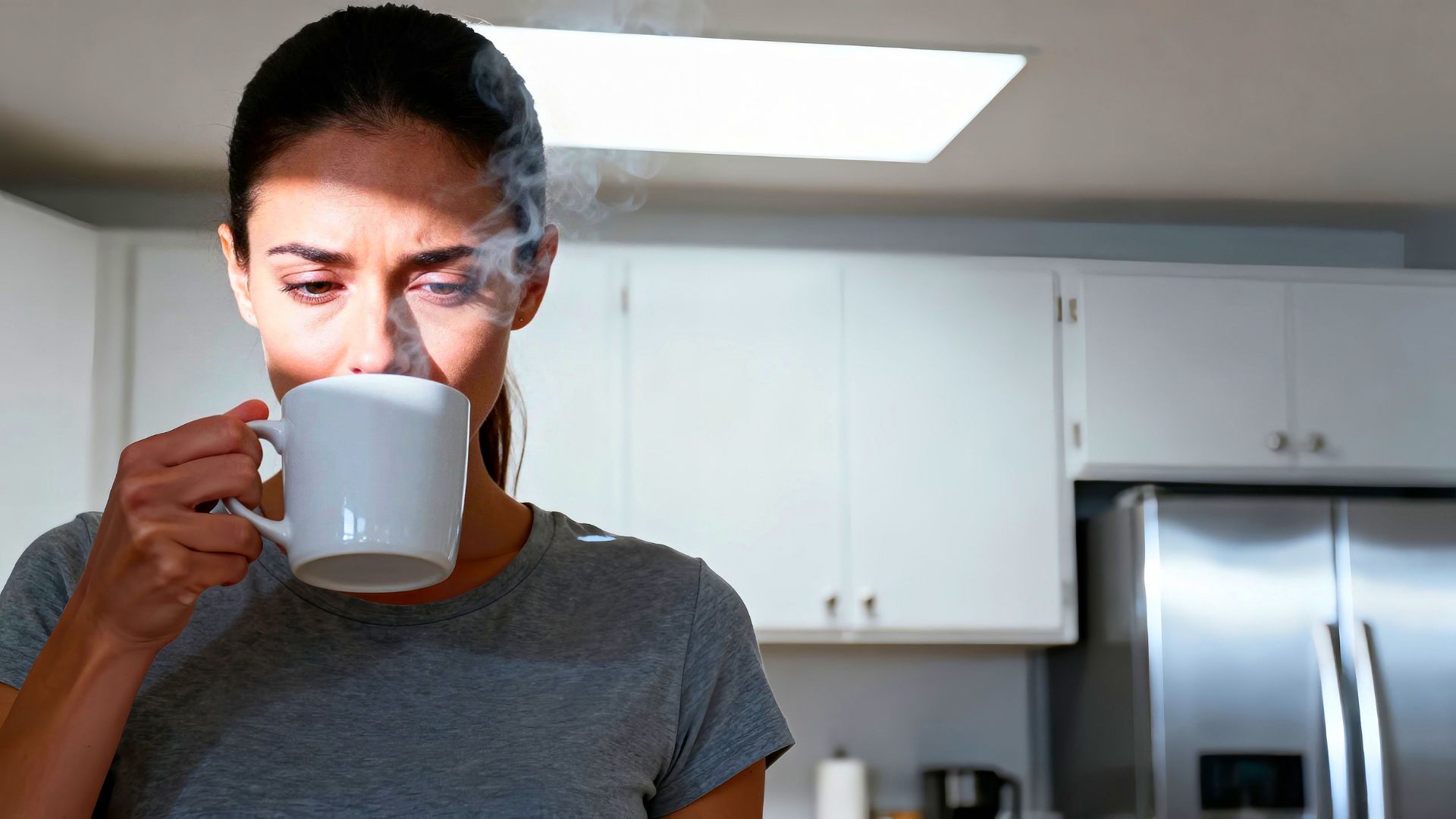A woman standing in a kitchen, holding a white mug close to her face as steam rises from the drink